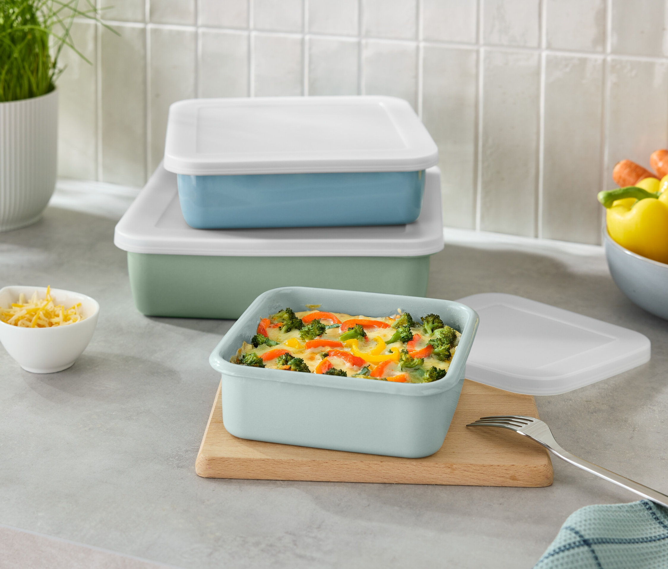 Egymásra rakott zöld és kék téglalap alakú tárolóedények fehér fedőkkel, valamint egy kis tál reszelt sajttal.