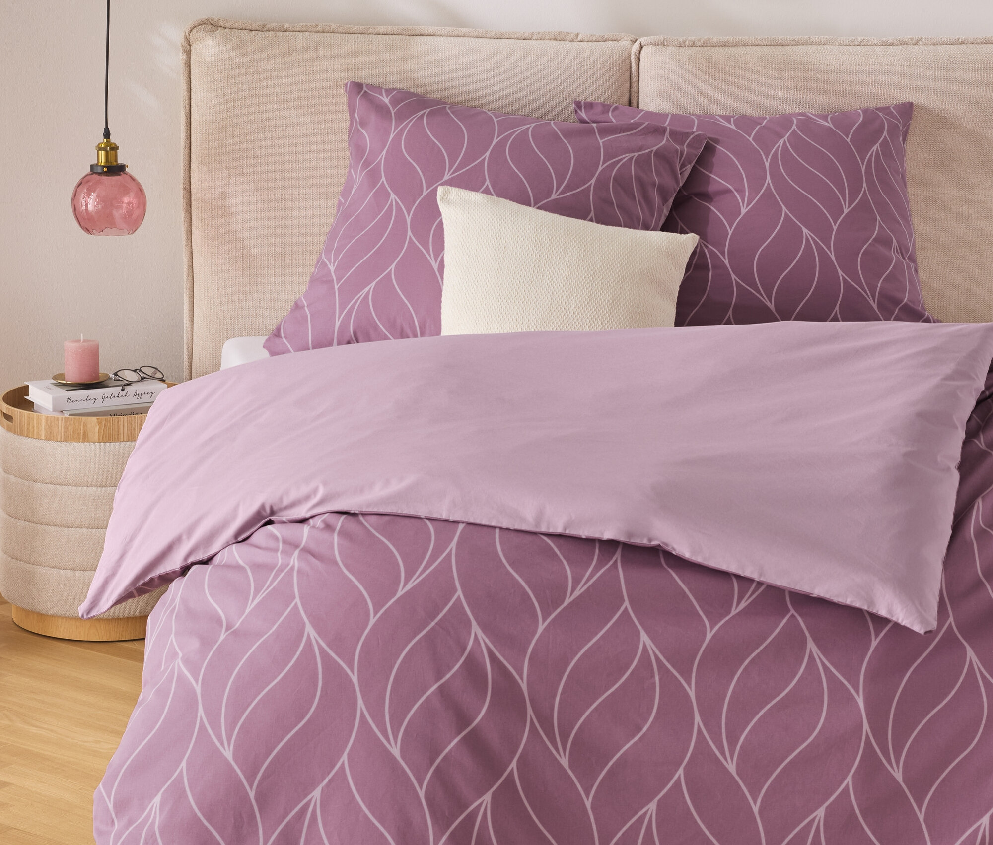 Lila ágynemű fehér levélmintával egy bézs fejtámlás ágyon. Az ágy mellett egy éjjeliszekrény áll lámpával.