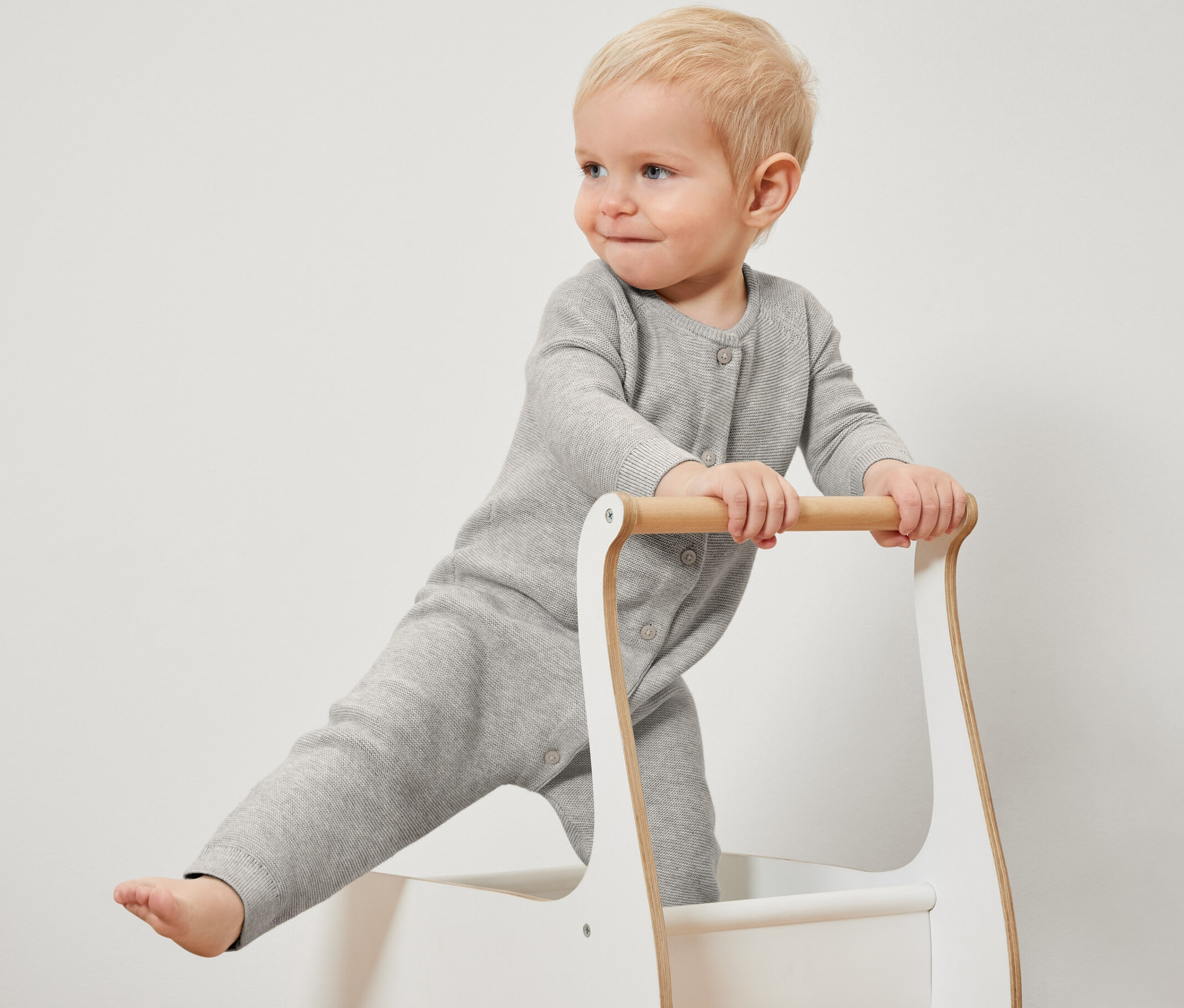 Egy szőke kisgyermek áll egy járgányban, szürke kötött kezeslábast visel.