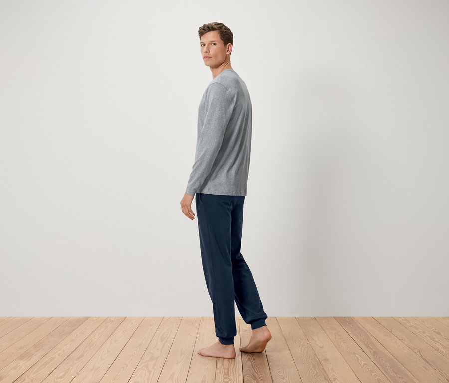 Szürke hosszú ujjú pólót és sötétkék nadrágot viselő férfi áll mezítláb a fapadlón, és a válla fölött néz hátra.