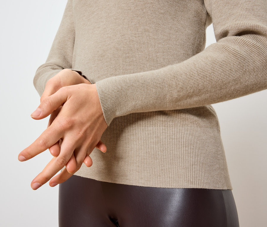 Nő dörzsöli a kezét egy krémszínű pulóver és barna leggings előtt.