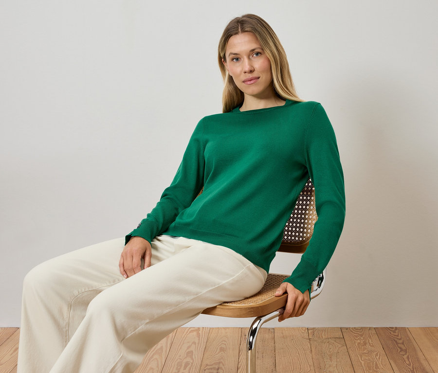 Egy szőke nő ül egy széken, zöld pulóvert és krémszínű nadrágot visel.