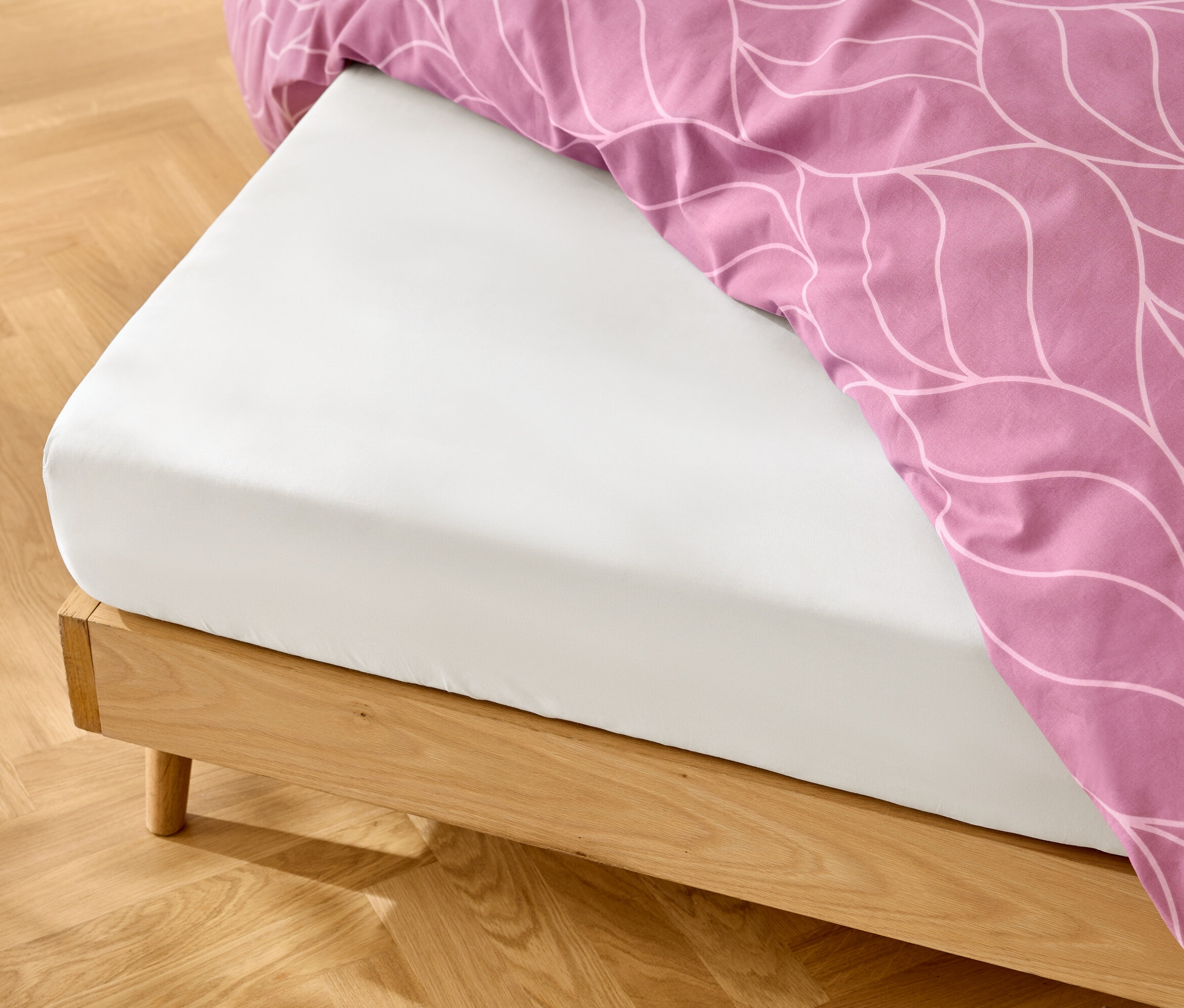 Fehér gumis lepedő fa ágykereten, rózsaszín ágynemű fehér mintával.