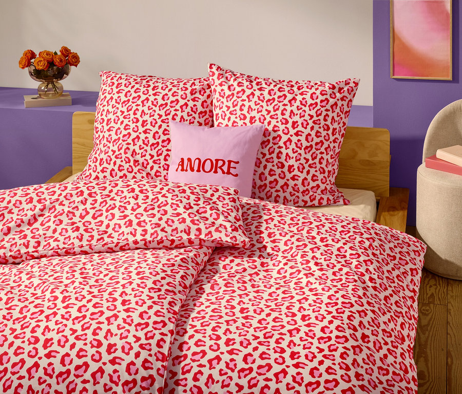 Egy ágy közeli képe piros-fehér leopárdmintás ágyneművel és párnákkal. Két párna előtt egy levendula színű párna látható "AMORE" felirattal.