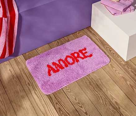 Egy lila fürdőszobai szőnyeg piros "AMORE" felirattal egy fa padlón fekszik egy lila éjjeliszekrény mellett.