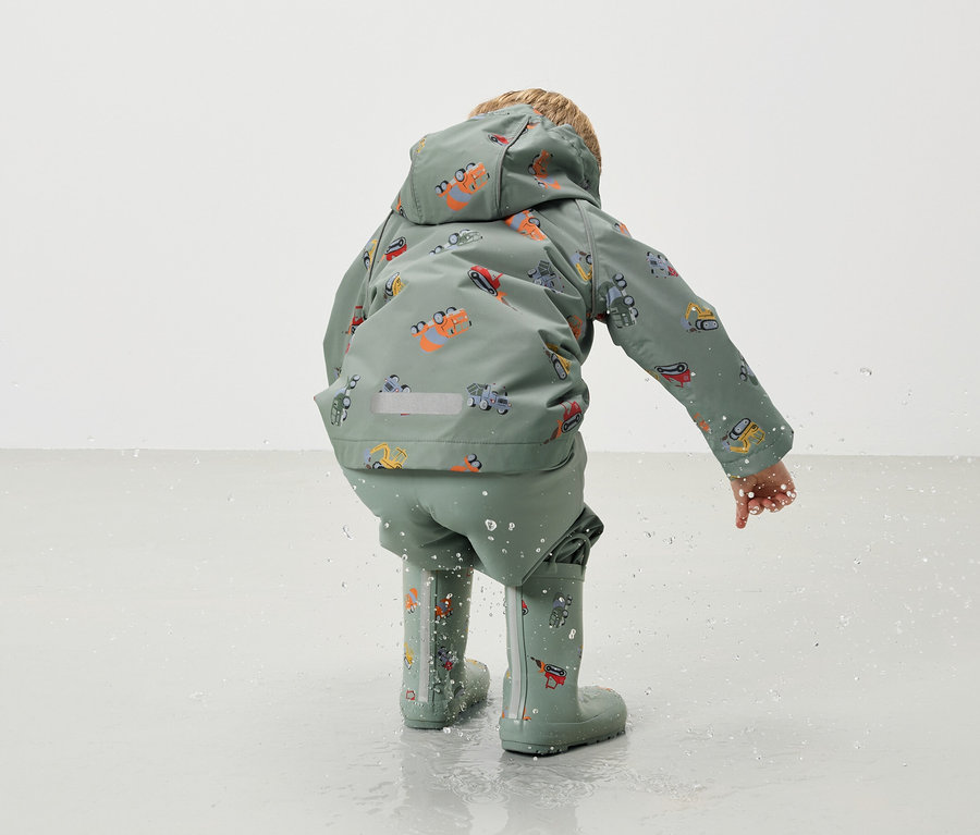 Egy hátulról látható gyermek zöld színű, markolómintás esőkabátot és esőnadrágot, valamint hozzáillő gumicsizmát visel, miközben a vízben játszik.