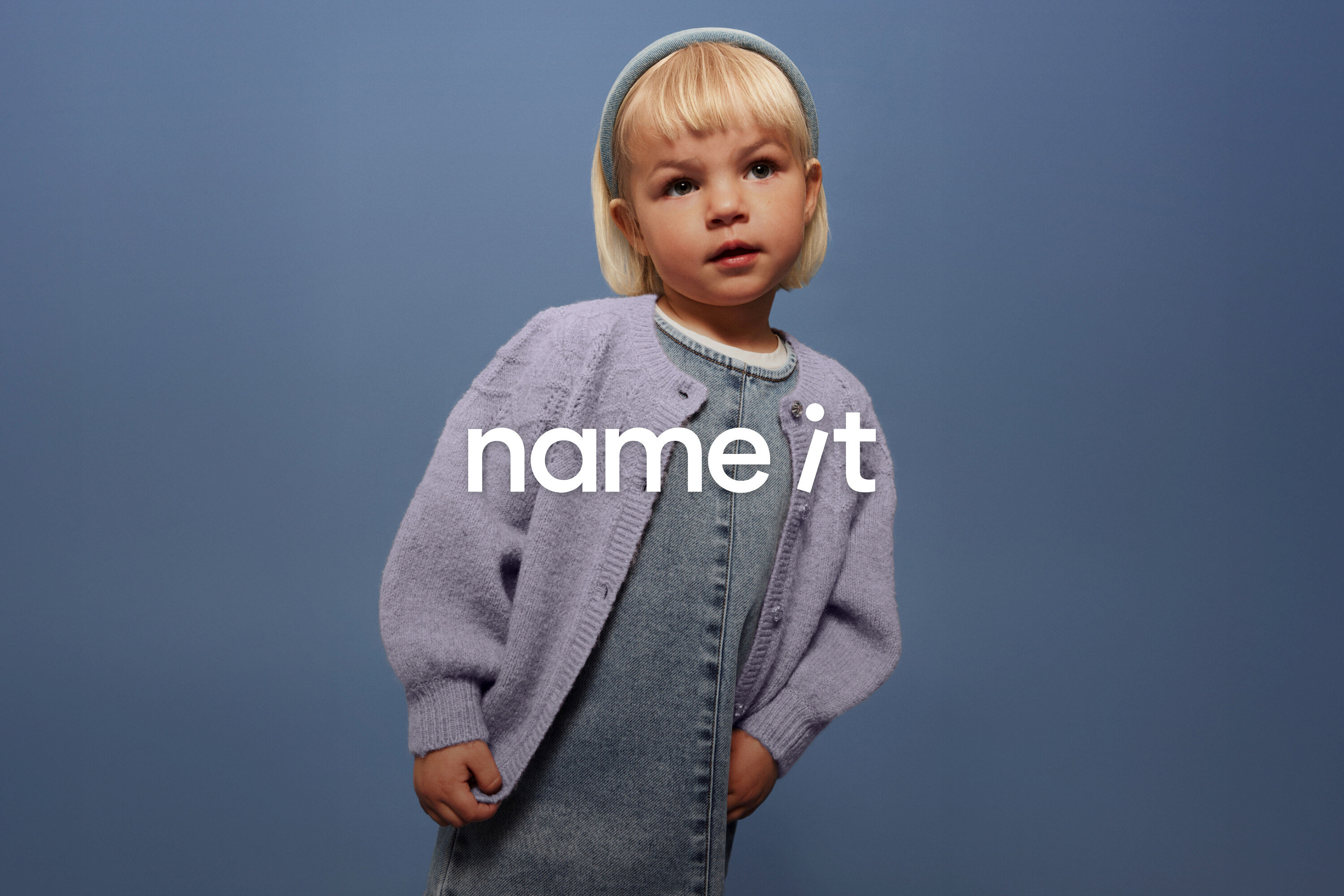 Egy szőke hajú, kék fejpántot viselő kisgyerek farmer kertésznadrágot és lila kardigánt visel egy kék háttér előtt.