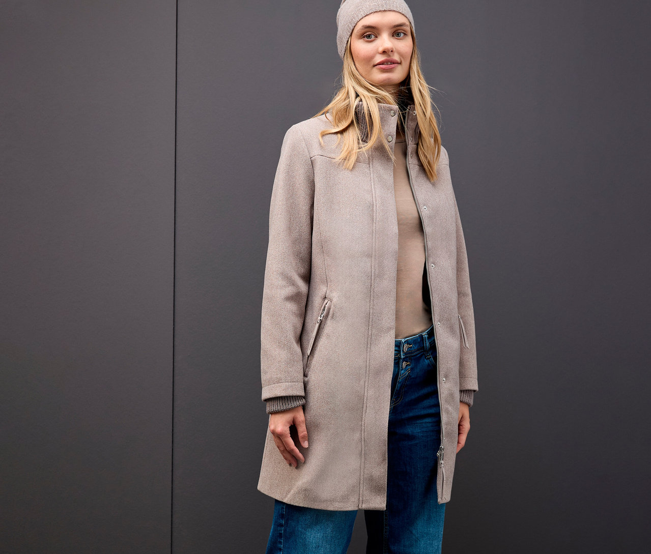 Egy szőke nő Street One rövid kabátot, sapkát, garbót és farmert visel szürke háttér előtt.