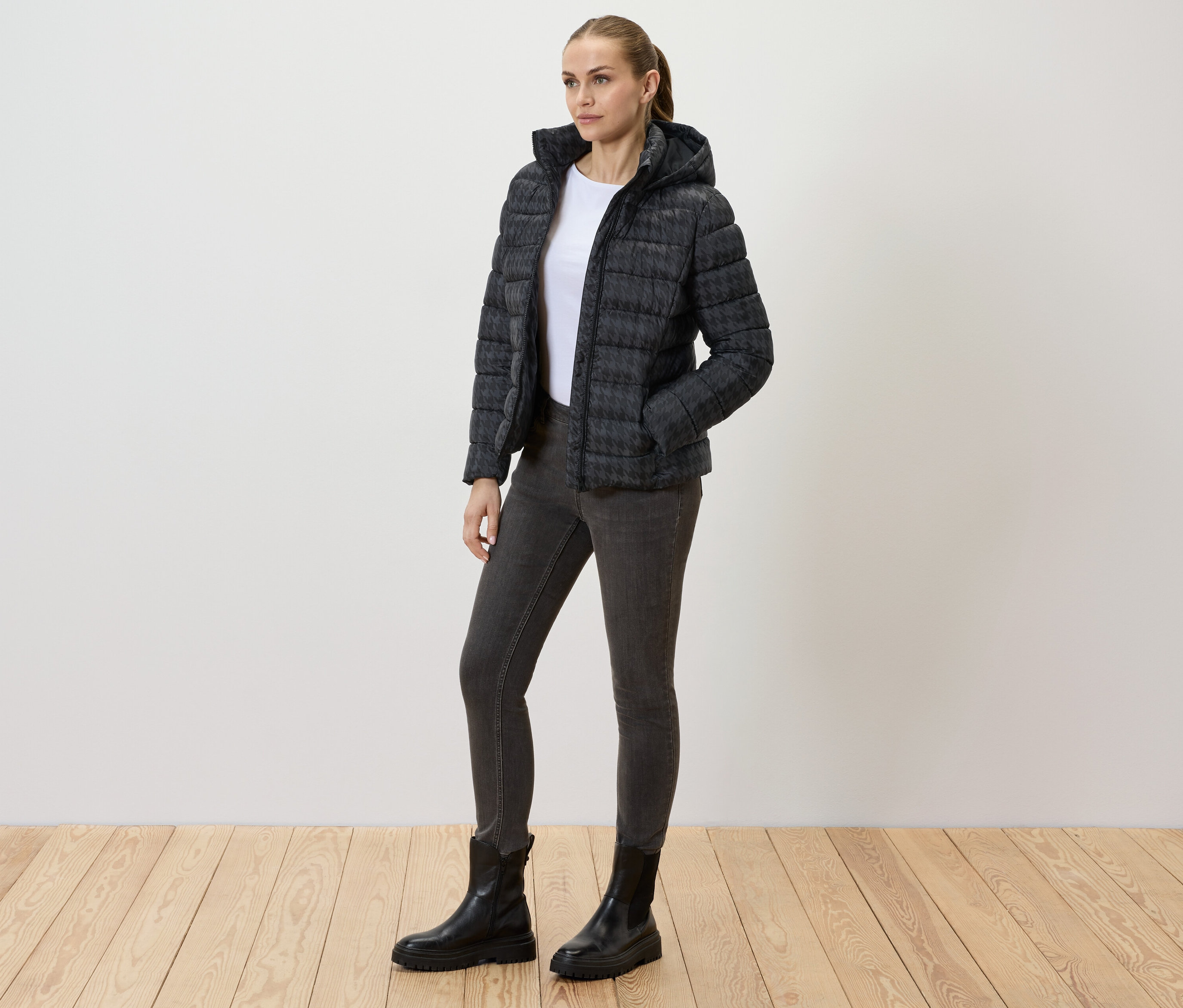 Nő visel nyomott mintás, kapucnis steppelt kabátot, farmert és fekete csizmát.