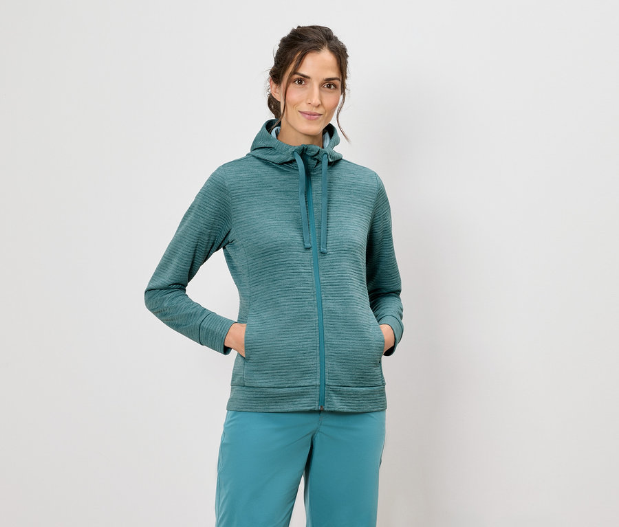 Nő kapucnis pulóverben és kék nadrágban áll fehér háttér előtt.