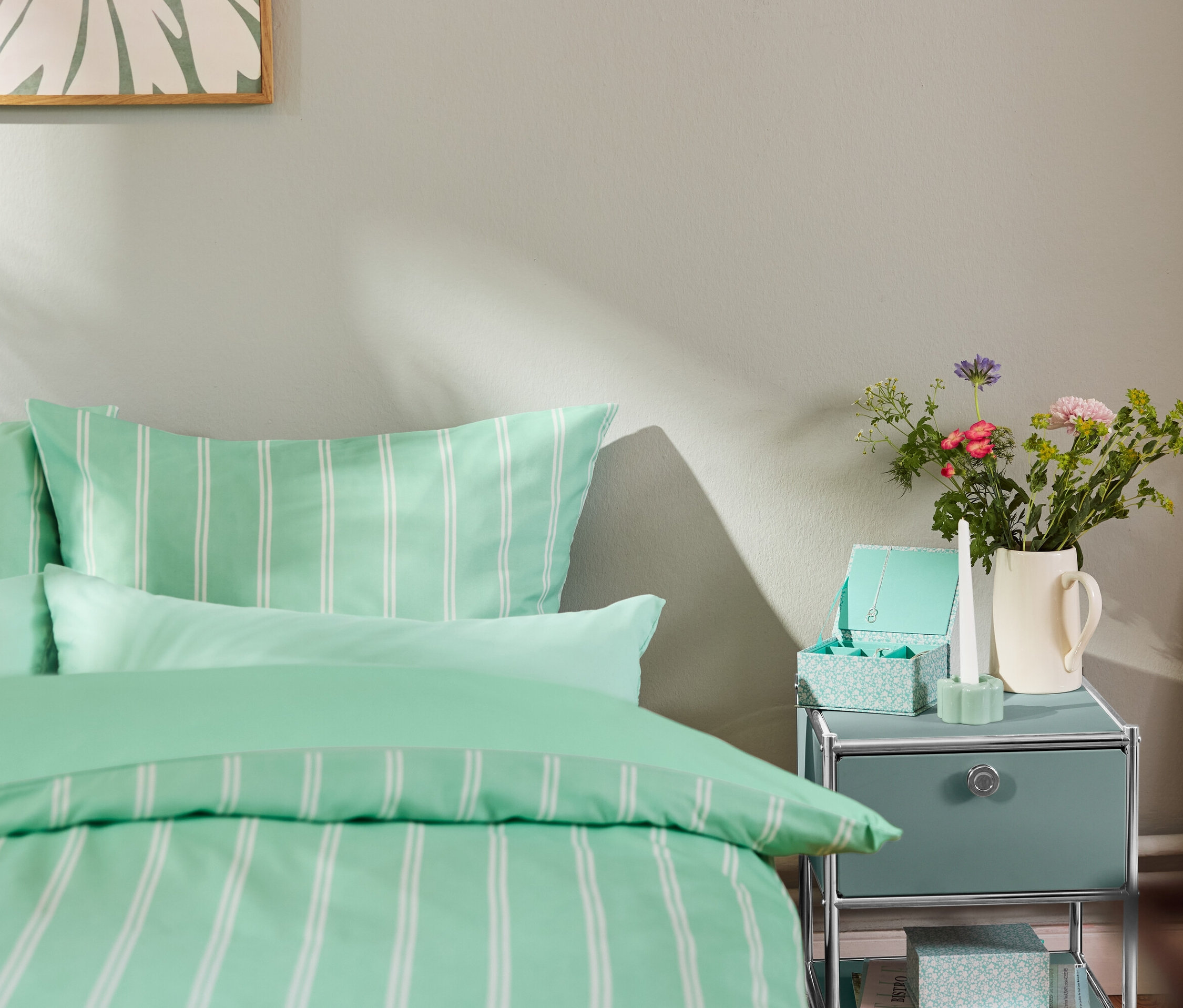 Ágynemű és zsályaszínű »CN3« fiókos fém kisasztal az ágy mellett.