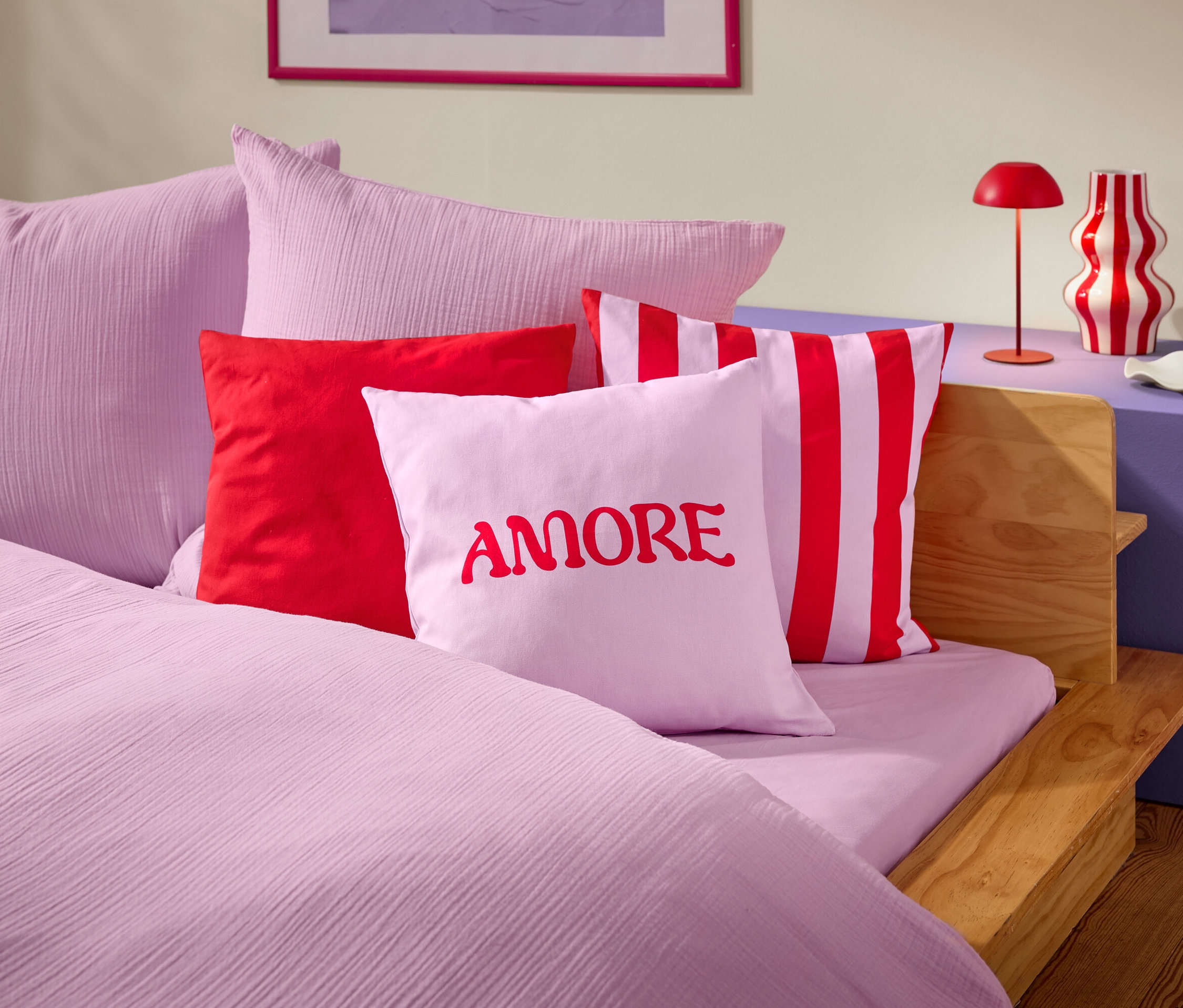Egy ágy közeli képe rózsaszín ágyneművel és párnákkal. Az egyik párnán piros betűkkel az AMORE szó látható.