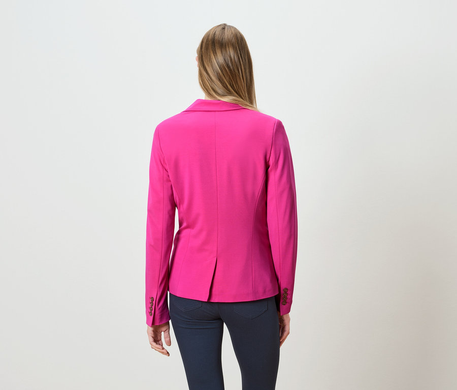 Hátulnézet egy hosszú szőke hajú nőről, aki rózsaszín blézert és sötét farmert visel.