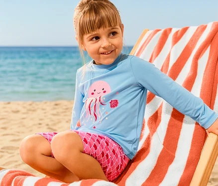 Egy kislány ül egy XL fürdőlepedővel ellátott nyugágyon, és Kinder-Kombi mit UV-Schutzfaktor 50+, Qualle szettet visel.