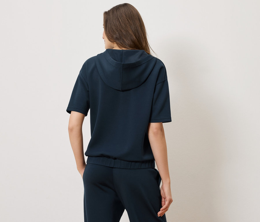 Hátulnézet egy nőről, aki sötétkék, rövid ujjú kapucnis pulóvert és hozzáillő nadrágot visel.