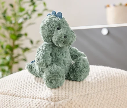 Egy zöld plüss dinoszaurusz ül egy fehér, texturált puffon. A háttérben egy növény és egy szekrény látható.