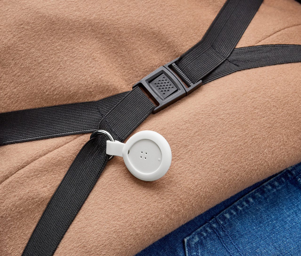 Egy fehér, kör alakú eszköz három ponttal egy fekete pántra van rögzítve egy fémgyűrűvel. A fekete műanyag csattal ellátott pánt világosbarna anyagon és kék farmeranyagon fekszik.