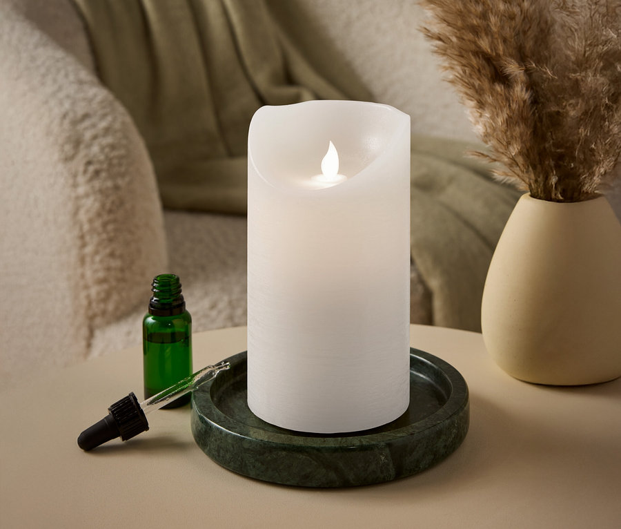 Egy fehér, LED lánggal égő gyertya áll egy zöld márványtálcán egy zöld üveg pipettás üveg mellett.