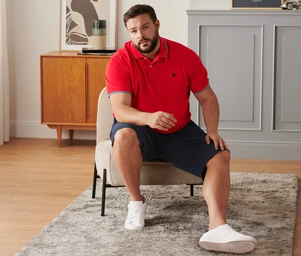 Egy férfi ül egy széken, tengerészkék chino rövidnadrágot, piros piké pólóinget és sneaker cipőt visel.