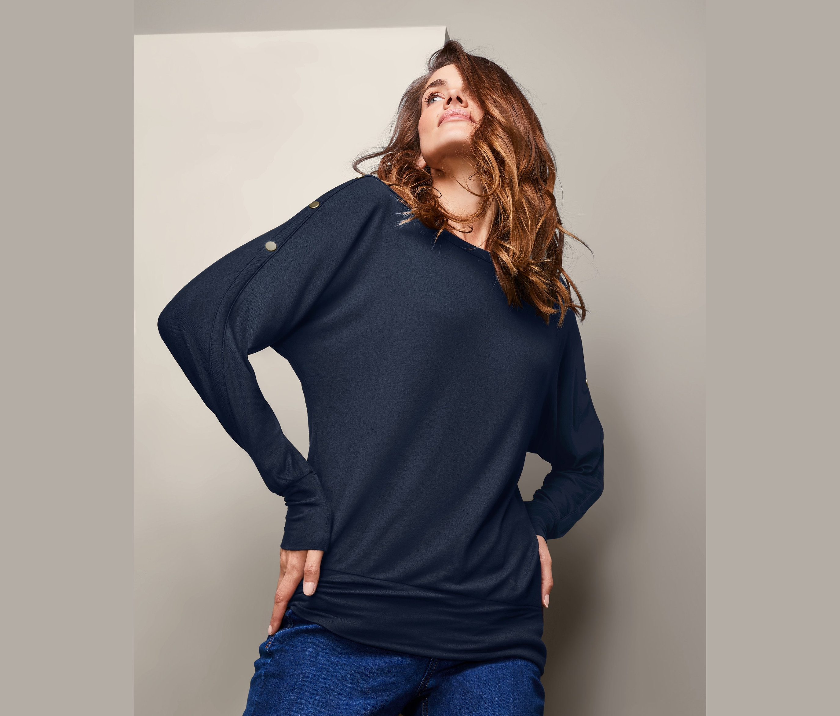 Nő pózol sötétkék, denevérujjú felsőben és farmerban.