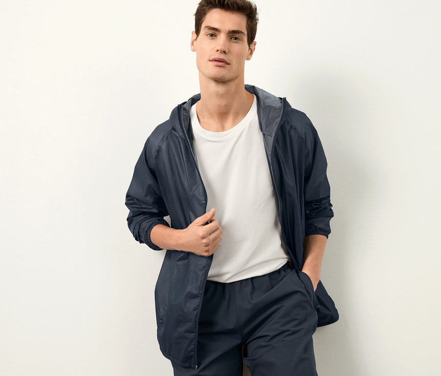 Férfi pózol a falnak dőlve, széldzsekit és rövidnadrágot visel.