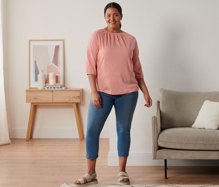 Egy nő áll és bőr szandált, 7/8-os treggingst és korall színű felsőt visel.