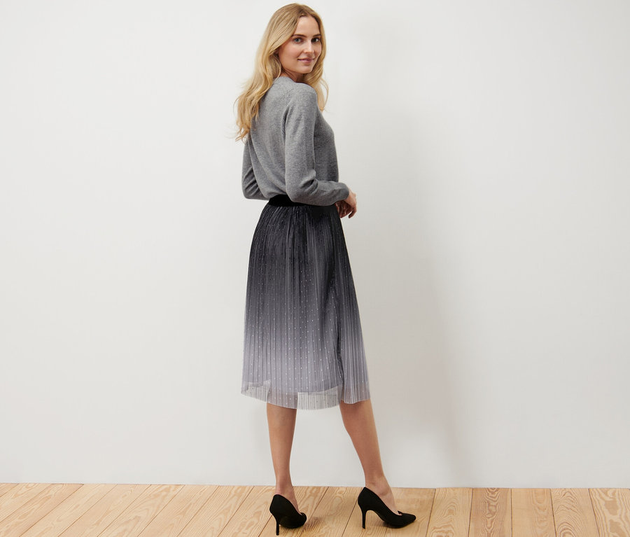 Nő pózol, pliszírozott, színátmenetes tüllszoknyát, szürke pulóvert és fekete magassarkút visel.