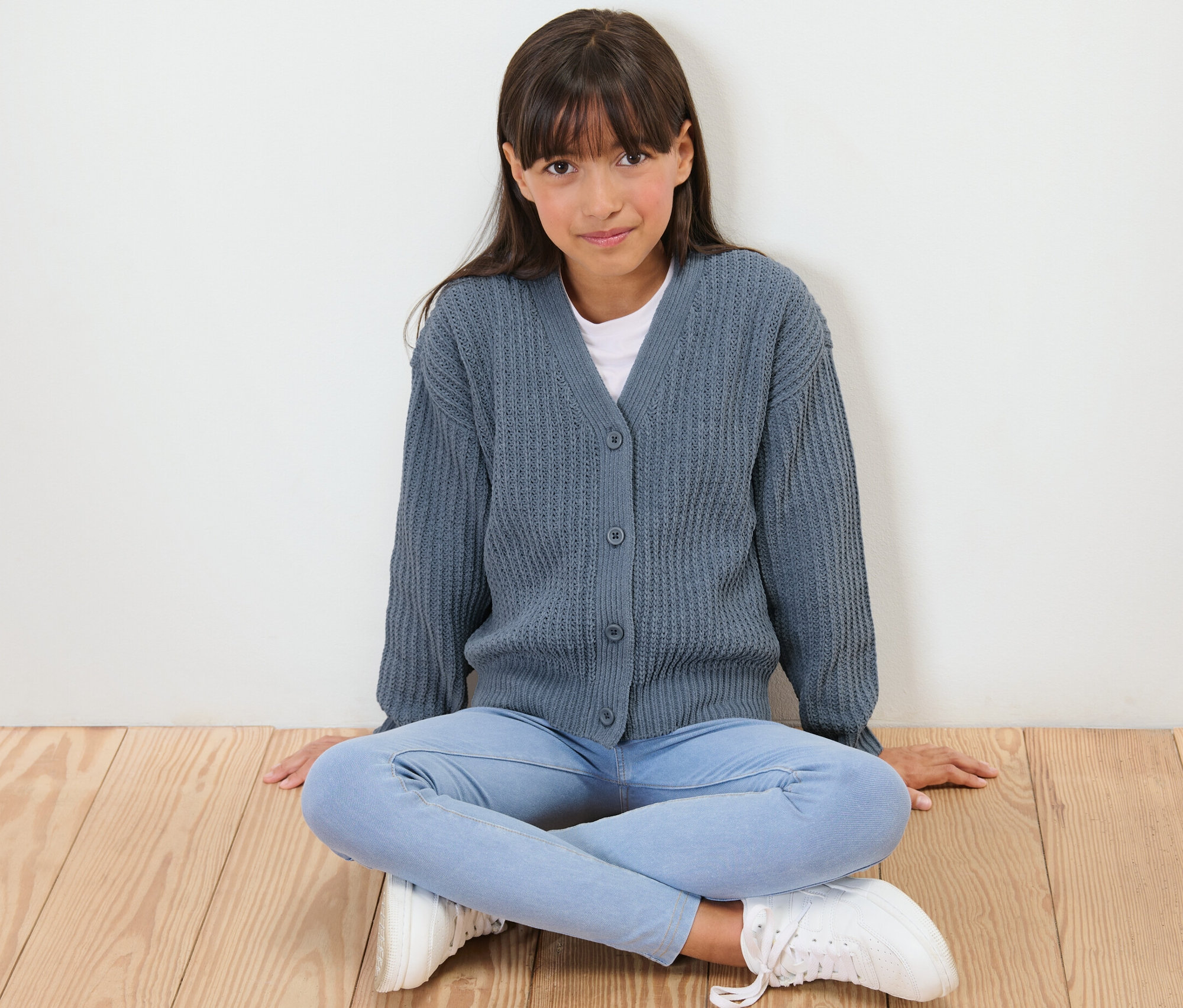 Lány ül a padlón, kék gyerek zsenília kardigánt visel.
