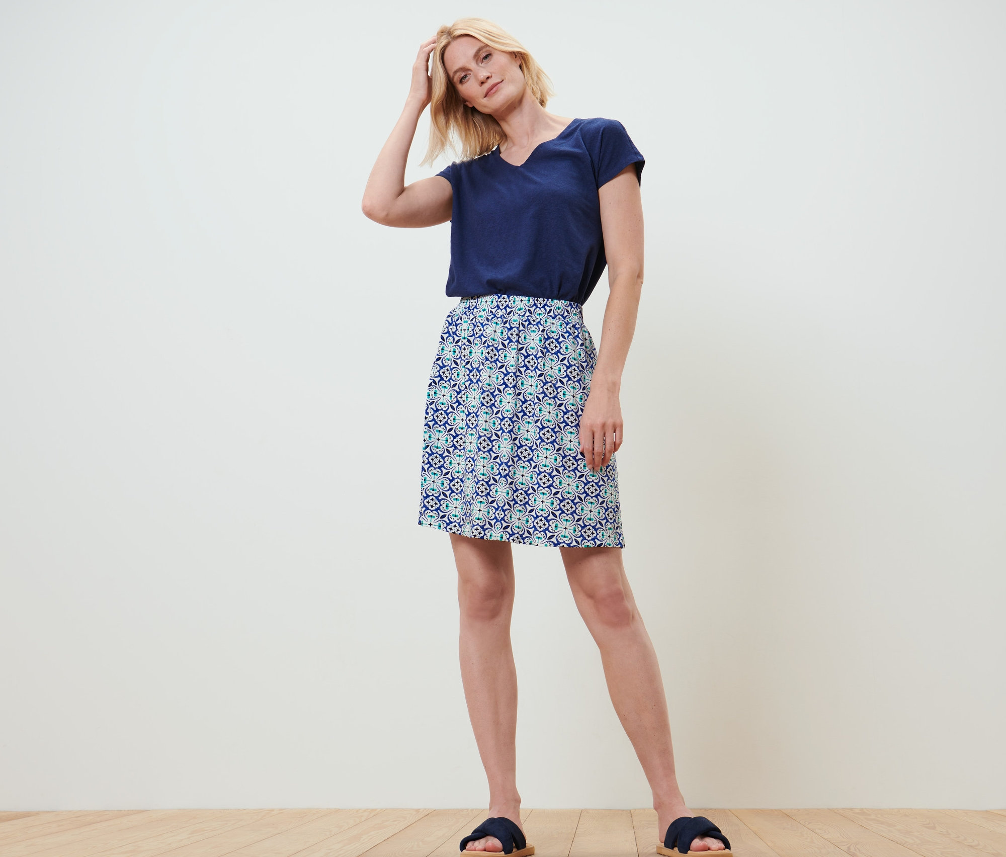 Egy szőke nő áll, kezével a hajában. Sötétkék pólót és nyomott mintás jersey szoknyát visel.