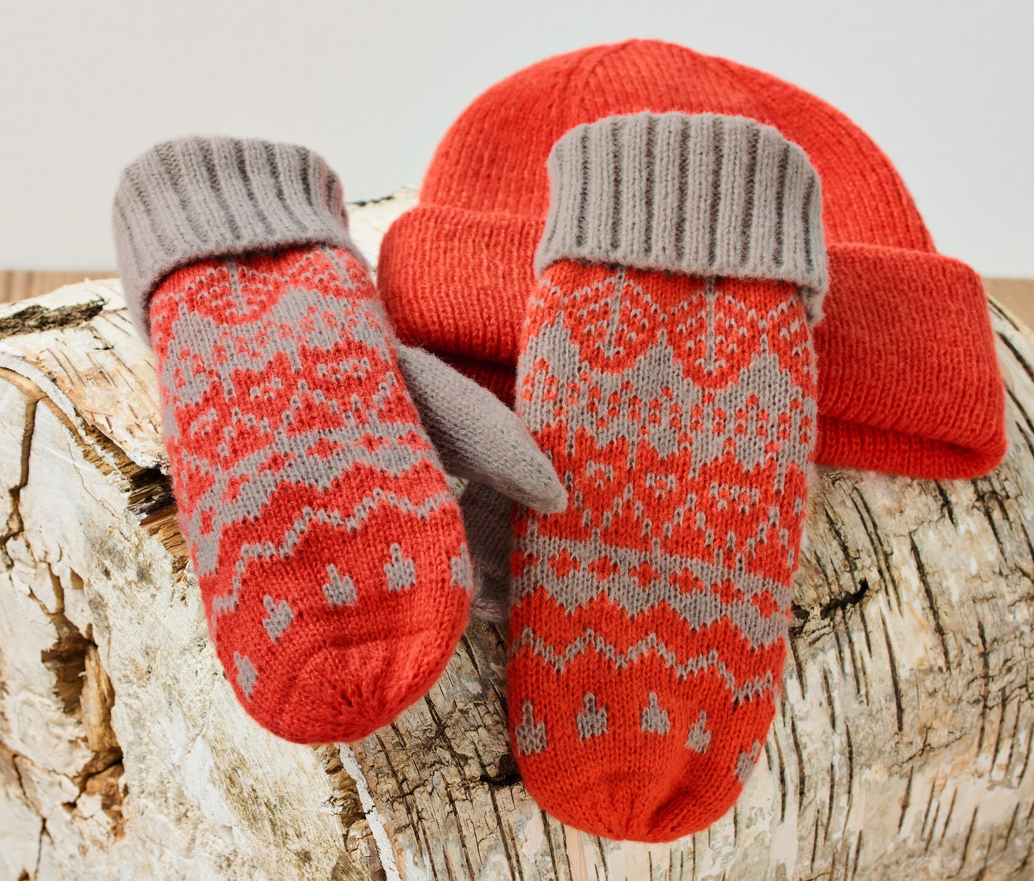 Kötött, norvég mintás női egyujjas kesztyű és narancssárga kötött sapka egy nyírfahasábon.