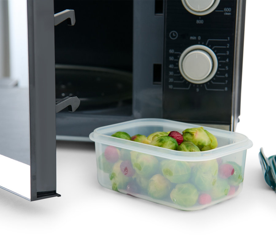 Egy nyitott mikrohullámú sütő, benne egy 3 darabos frissentartó doboz rózsákkal és málnával. A doboz mellett egy zöld fedő található.