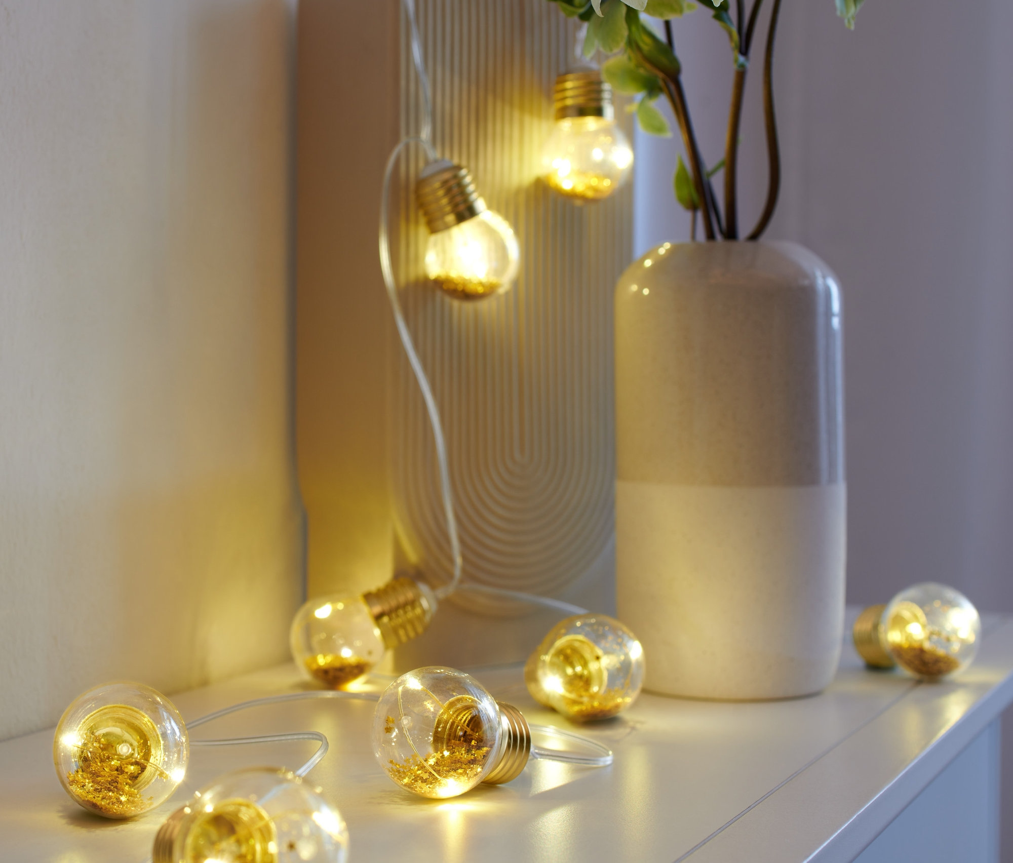 Egy LED-es villanykörtés fényfüzér egy fehér polcon fekszik egy váza és egy képkeret mellett.