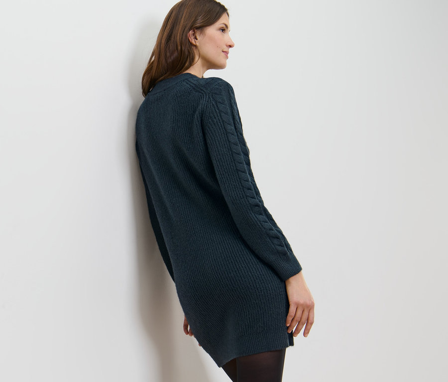 Egy nő egy fehér falnak dőlve sötétkék kötött ruhát visel.