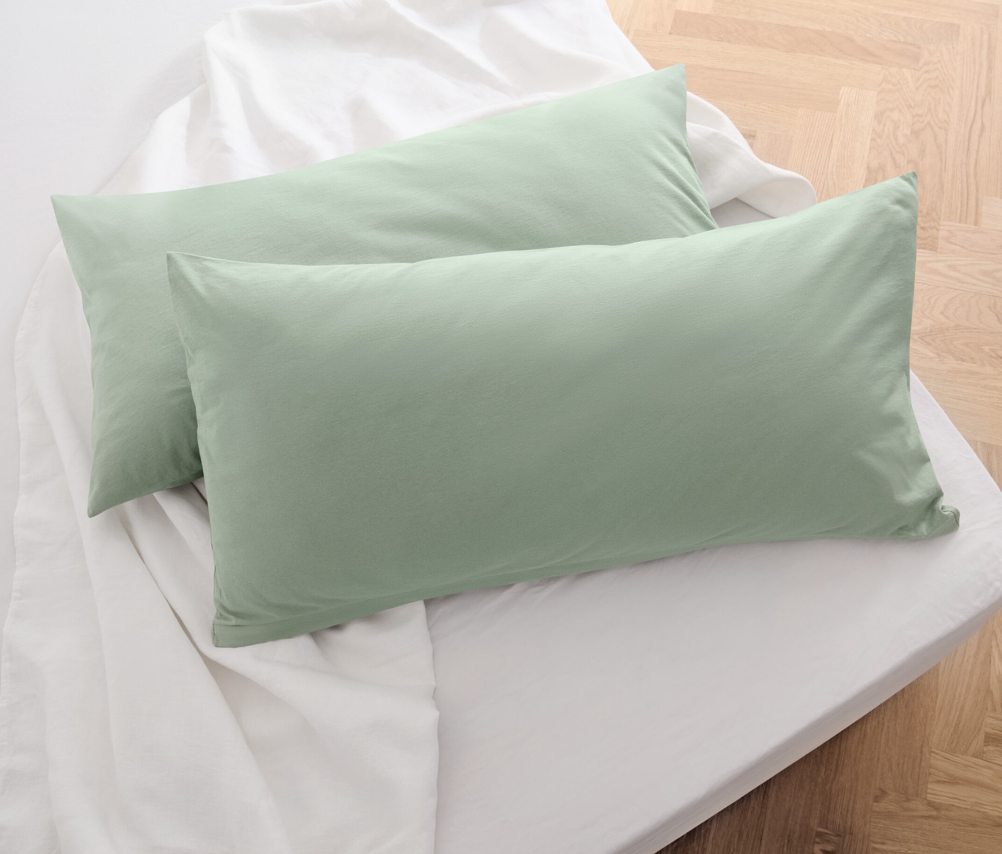 Két zsályaszínű jersey párnahuzat fekszik egy fehér lepedőn.