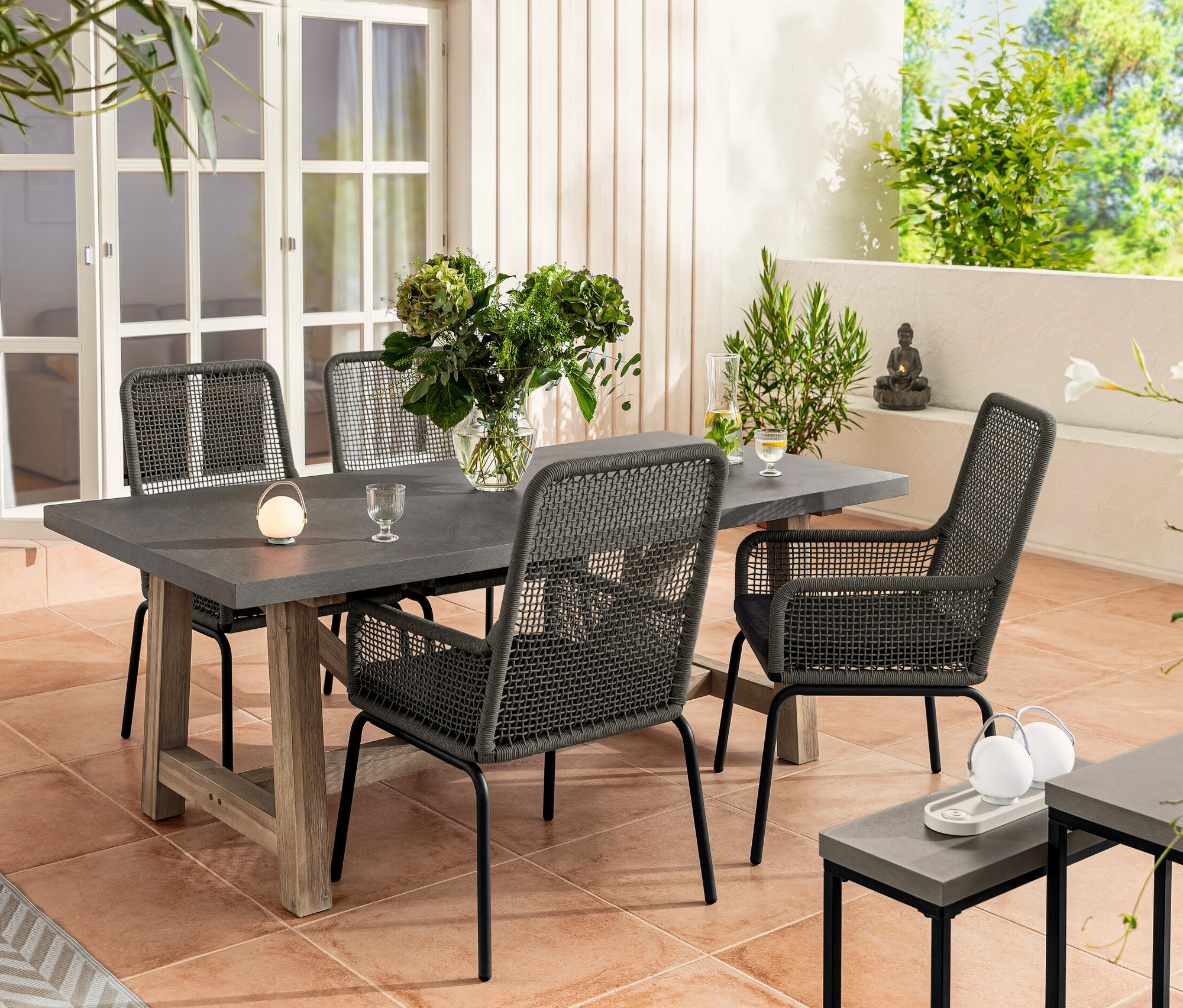 Négy textilhálós, magas háttámlás szék áll egy policement asztallapos étkezőasztalnál. Az asztalon egy váza virág és egy akkumulátoros asztali lámpa szett található. A háttérben egy kert és egy konzolasztal-szett, policement asztallappal látható.