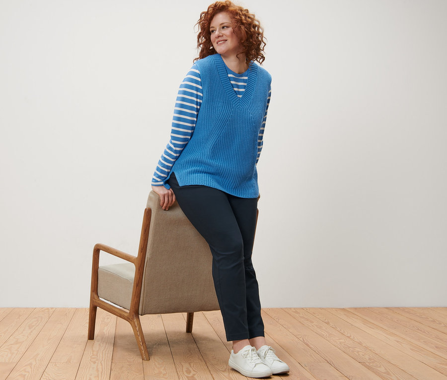 Egy göndör, vörös hajú nő egy széknek támaszkodik, csíkos hosszú ujjú felsőt, kötött mellényt és sötétkék női bengalin nadrágot visel.