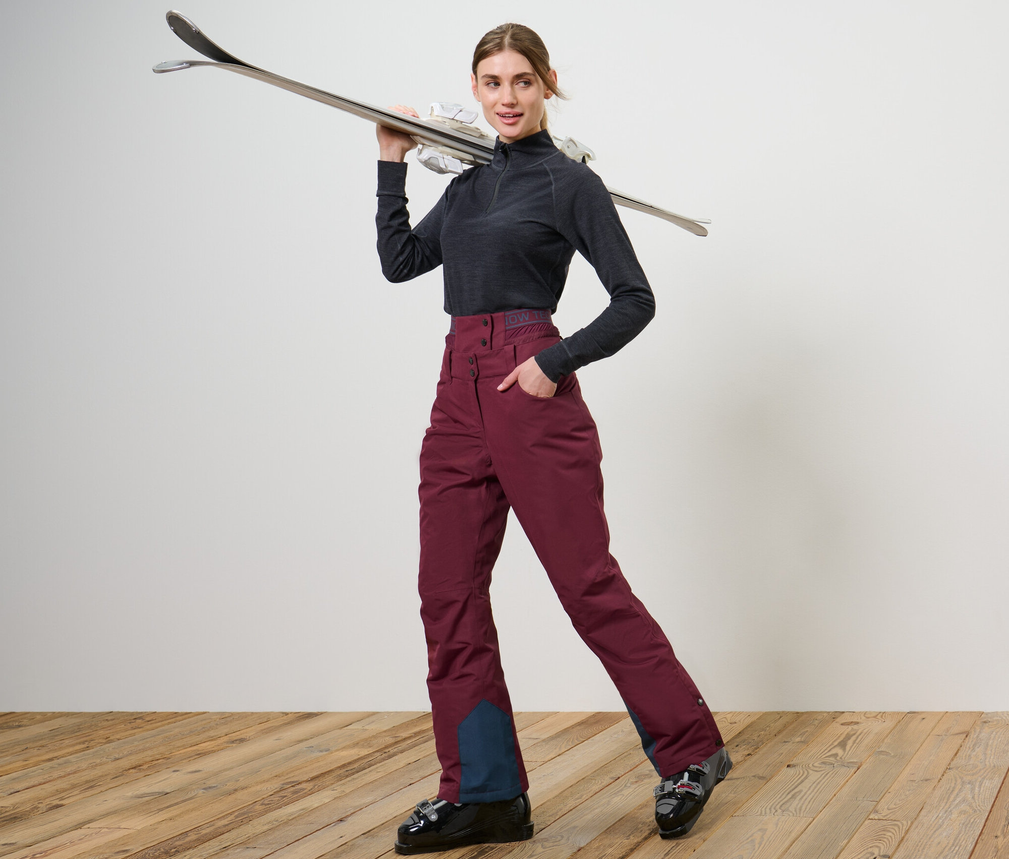 Nő pózol síbakancsban és borvörös sí- és snowboardnadrágban.