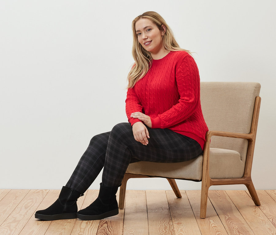 Nő ül egy fotelben, piros fonott mintás kötött pulóvert és kockás női lezser nadrágot visel.
