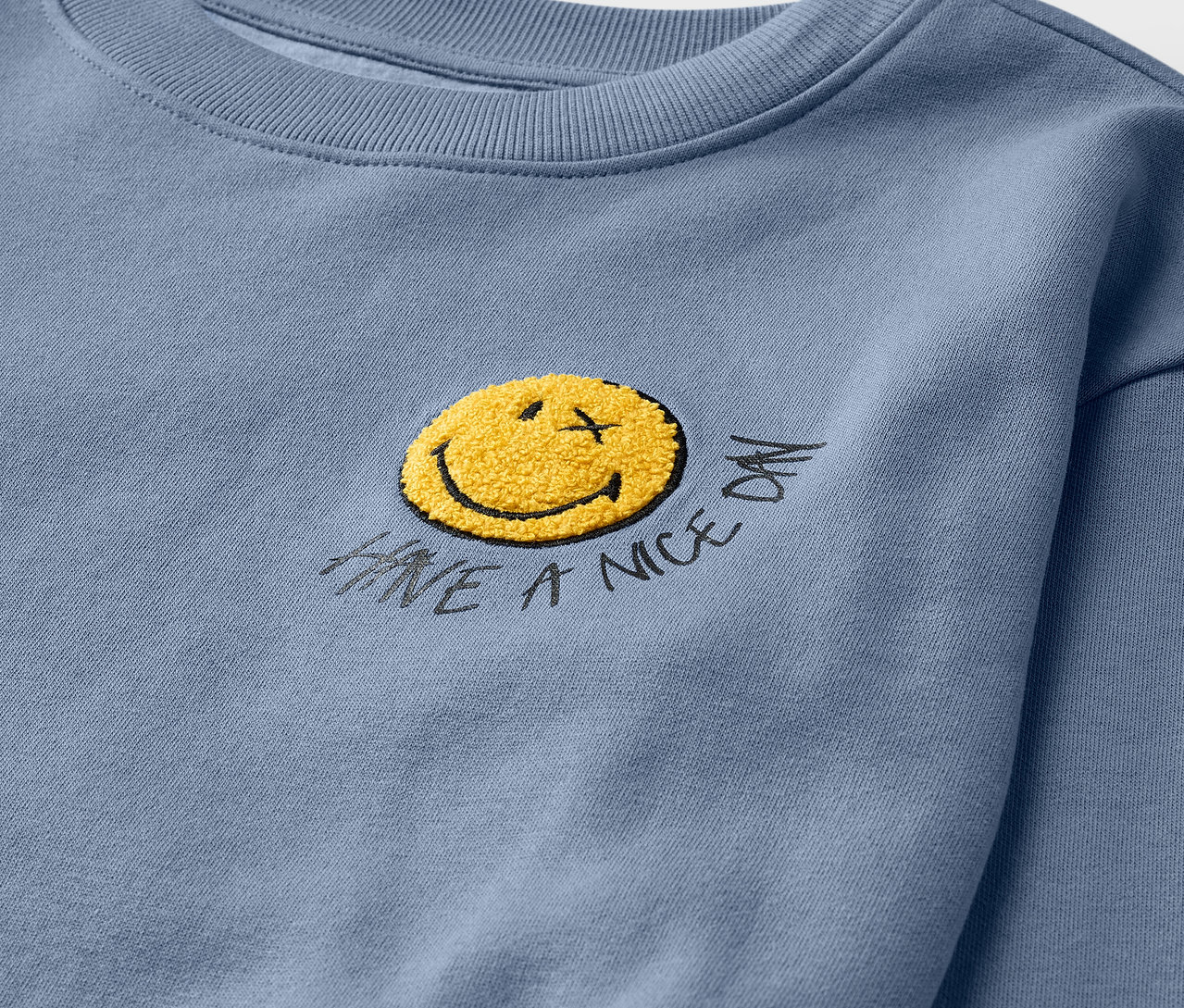 Egy világoskék SMILEYWORLD® gyerek szabadidőfelső részletének közeli képe, bolyhozott belsővel és egy sárga mosolygós arccal.