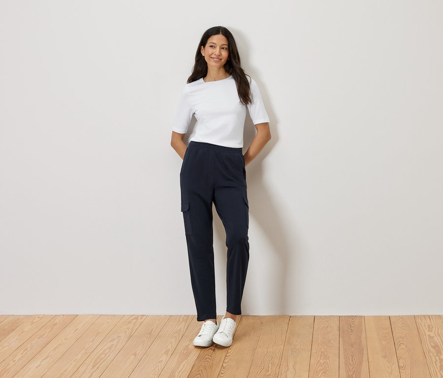 Nő áll összefont karokkal a fal előtt, fehér pólót, sötétkék cargo nadrágot és fehér tornacipőt visel.