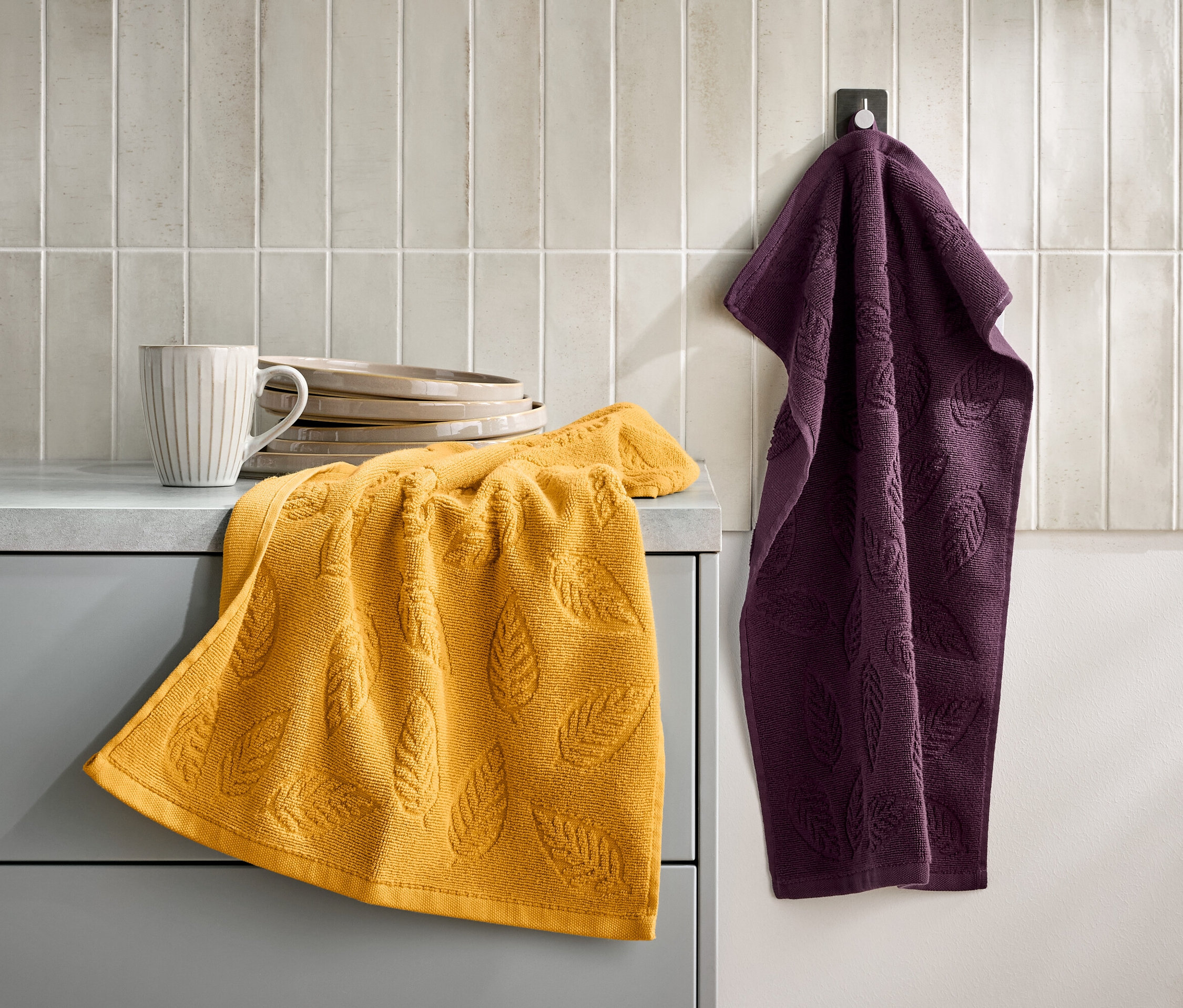 Egy levélmintás sárga törölköző egy szürke szekrényen fekszik egy csésze és tányérok mellett. Egy lila törölköző lóg egy kampón a falon.