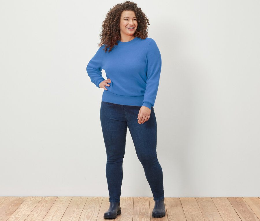 Nő pózol a fapadlón kék, finomkötésű pulóverben és farmerleggingsben.