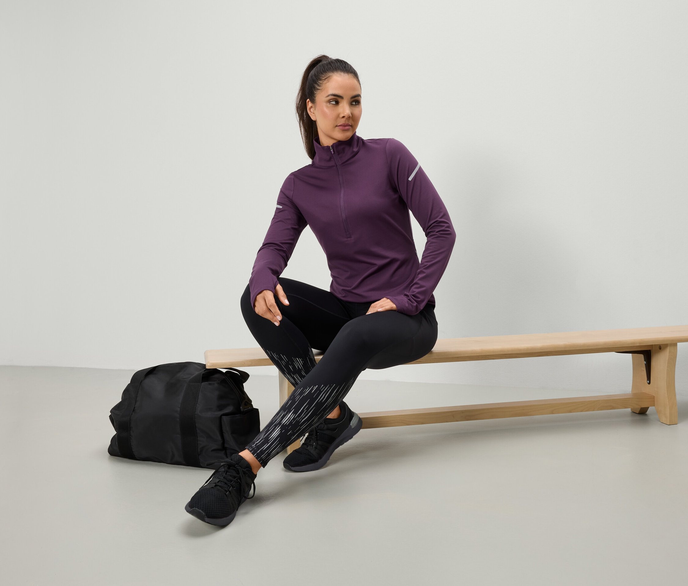 Egy nő lila sportfelsőben és fekete leggingsben ül egy padon, mellette egy fekete sporttáska.