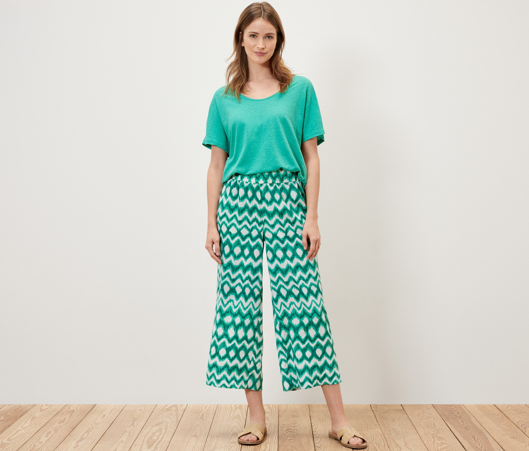 Egy nő áll zöld pólóban és nyomott mintás szatén nadrágban.