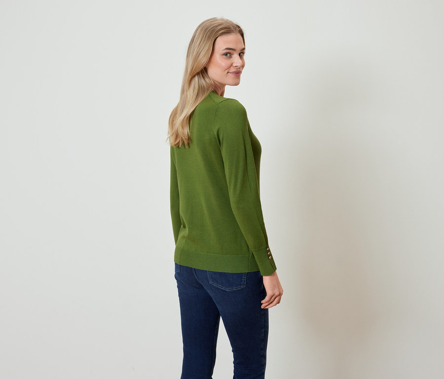 Egy nő visszanéz a válla fölött, olívazöld pulóvert és sötétkék farmert visel.
