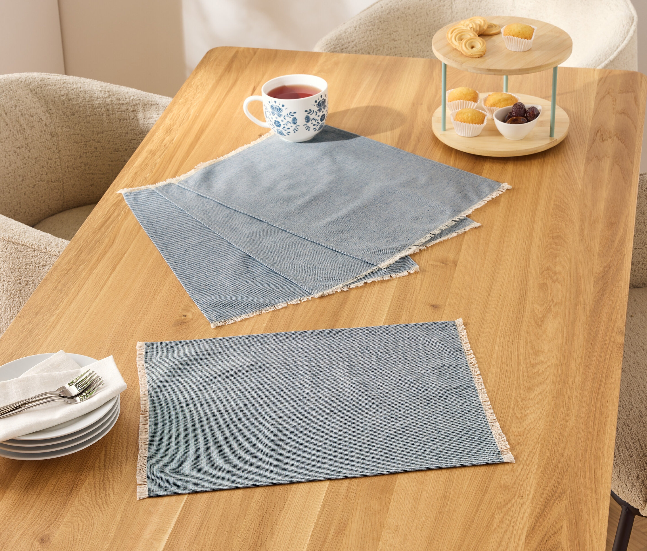 Két kék, fehér szegélyű teríték egy fa asztalon, mellette egy csésze, tányérok és egy emeletes tál étellel.