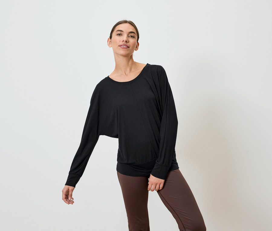Nő fekete, hosszú ujjú felsőben és barna leggingsben pózol fehér háttér előtt.