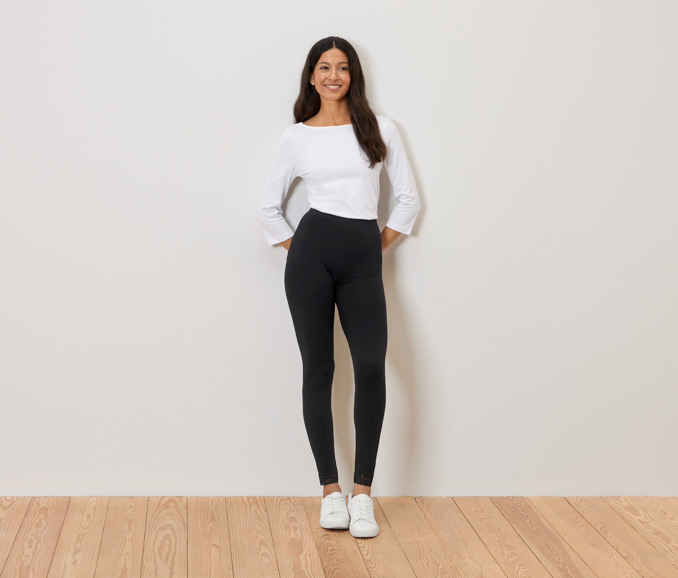 Nő áll a fal előtt, fehér felsőt, fekete leggingset és fehér tornacipőt visel.