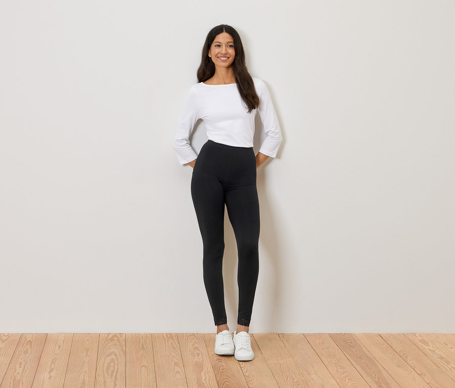 Nő áll a fal előtt, fehér felsőt, fekete leggingset és fehér tornacipőt visel.