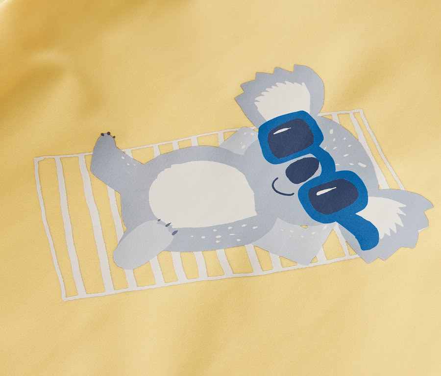 Sárga gyerekpóló UV-faktorral 80, koala kék napszemüvegben fekszik.